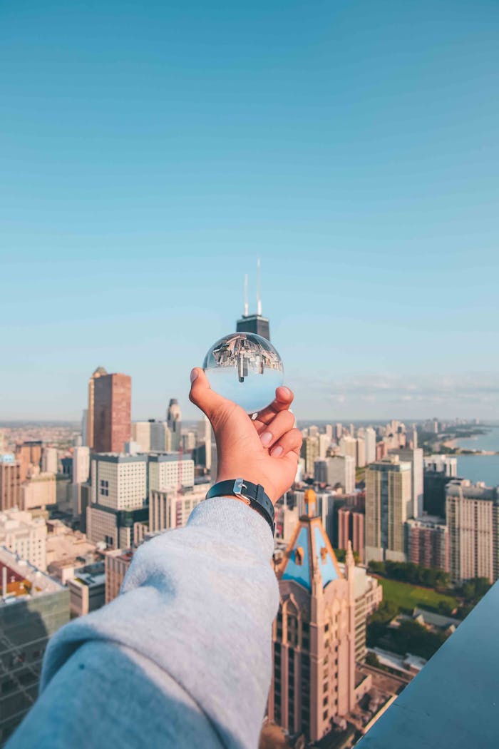 A hand holds a glass sphere reflecting a city's skyline on a sunny day.